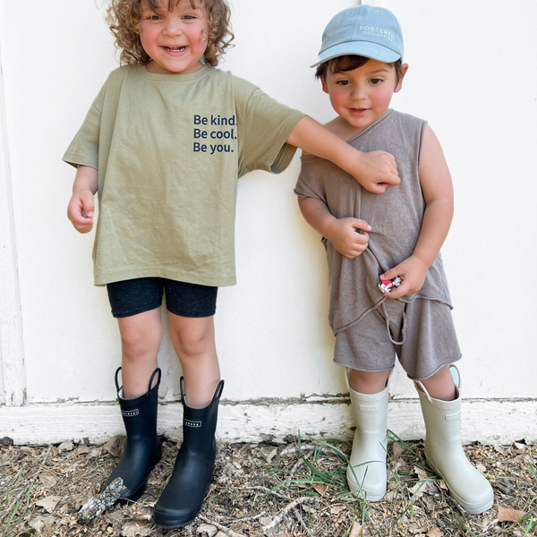 Lifestyle images of two boys in Denver James Rain boots next to a wall in the dirt laughing. One boy is wearing black puddle buddies and the other is wearing sand color rain boots in the same solid color way.