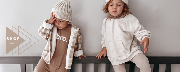 TWO BOYS, ONE WEARING BROWN ROMPER THAT SAYS BRO WITH CREAM BEANIE. OTHER WITH CREAM SWEATSHIRT AND BEANIE