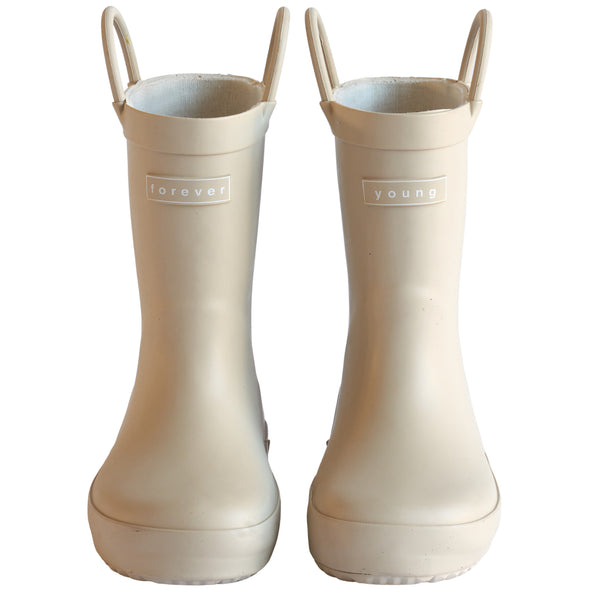 Front view of a pair of sand colored rain boots for children. Showing pull handles for easy slip on and tags that say "Forever Young" on the front.
