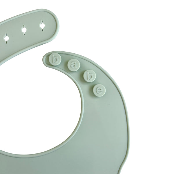 Close-up of an light green silicone bib with buttons labeled 'babe' on a white background