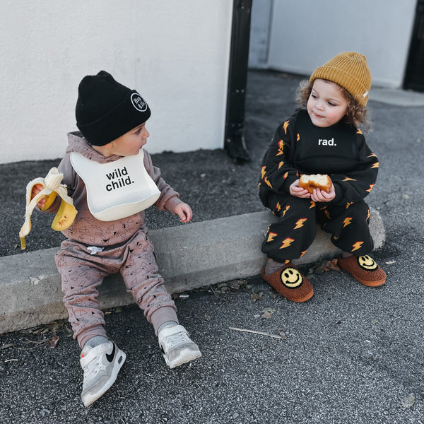 lifestyle image of 2 kids sitting on a curb with silicone bibs on on that says 'wild child.' and the other says 'rad.'