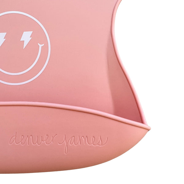 Pink silicone bib detail image with a smiley face design and 'denver james' branding on a white background