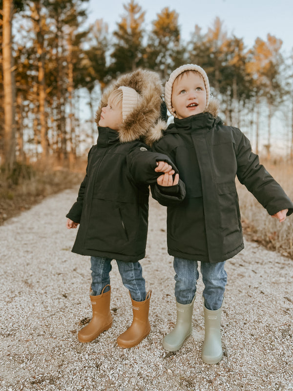 two children in rain coats holding hands on a gravel path in the woods both wearing jeans while wearing Denver James puddle buddies rain boots one in sand and the other in the golden colorway.