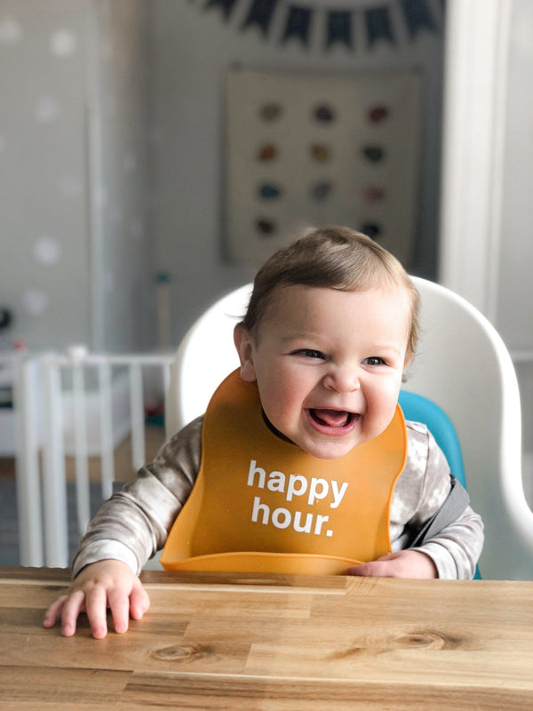 lifestyle image of baby boy wearing a silicone food catcher bib at a table with white text that says 'happy hour.'