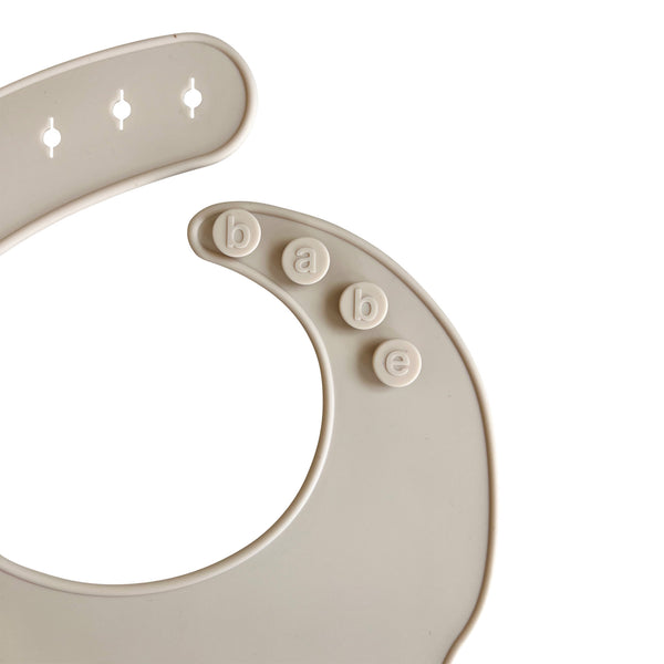 Close-up of an light grey silicone bib with buttons labeled 'babe' on a white background
