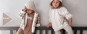 TWO BOYS, ONE WEARING BROWN ROMPER THAT SAYS BRO WITH CREAM BEANIE. OTHER WITH CREAM SWEATSHIRT AND BEANIE