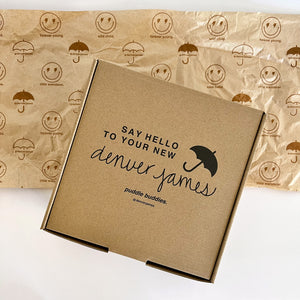 Branded Delivery box saying 'Denver James' on a white background with playful packaging paper with smily faces with lighting bolts for eyes and umbrellas. 
