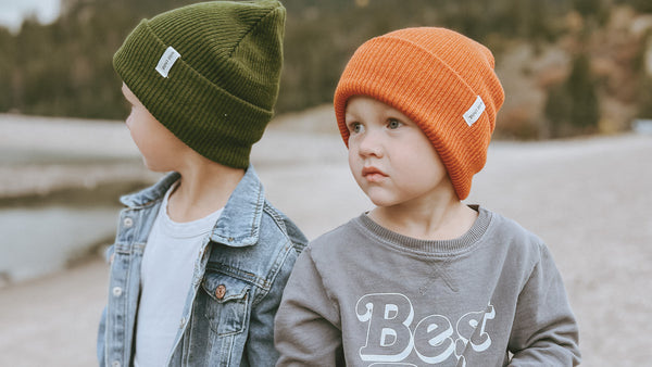Two children wearing green and orange beanies with visible brand logos, standing outdoors.