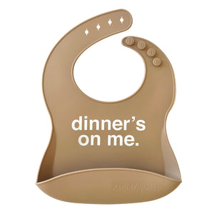  Front view of a beige silicone food catcher baby bib with 'dinner's on me' text on a white background