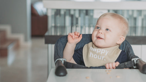 baby wearing bib that says 'happy hour' at kitchen table