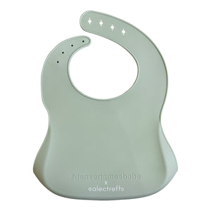 Back view of a light green silicone bib on a white background with '#denverjamesbabe' text and 'x @alectreffs' in white text. 