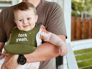 Baby wearing a bib with 'fork yeah' text, held by an adult outdoors.