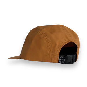 Back 3/4 view of a brown five-panel cap with a tag that has a smily face with lighting bolt eyes on a white backdrop. The hat has a one snap closure. 