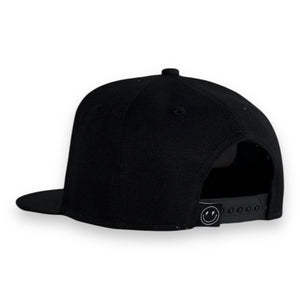 back 3/4 view of black 5 panel black baseball cap with a snap back and smiley face with lighting bolt eyes tag on white backdrop. 