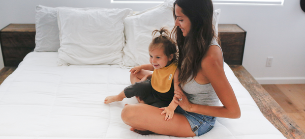 Mother and daughter sitting on bed, daughter is wearing a ribbed baby big in golden color while sitting on Mommys lap. 
