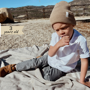 Light Brown | Kid sitting on pebble driveway wearing light brown beanie. 