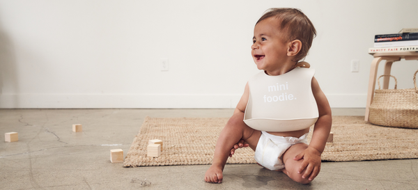 baby sitting on floor wearing 'mini foodie' silicone bib in a diaper. 