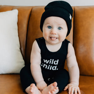 image of baby sitting on couch wearing black beanie and romper that says 'wild child'. 