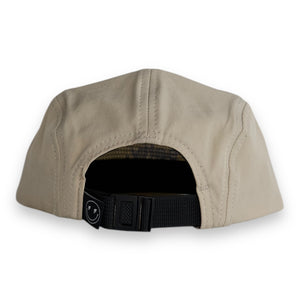 Back view of a tan five-panel cap with a tag that has a smily face with lighting bolt eyes on a white backdrop. The hat has a one snap closure. 