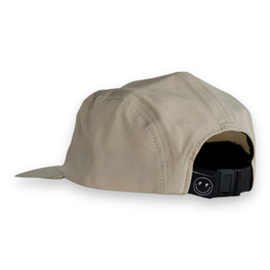 Back 3/4 view of a tan five-panel cap with a tag that has a smily face with lighting bolt eyes on a white backdrop. The hat has a one snap closure. 