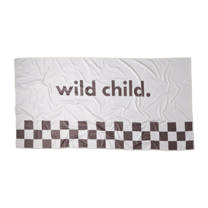 an overhead image of a stone or white colored microfiber beach towel with racing checkers along the bottom and the words 'wild child.' printed in large brown  letters across the middle on a flat surface showing the rectangular dimension of the towel. 
