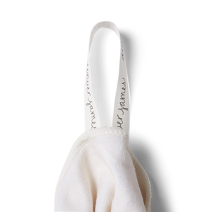 a detailed image of a hanging tag on a stone or white microfiber beach towel with 'denver james' printed in black cursive font on a white tag. 