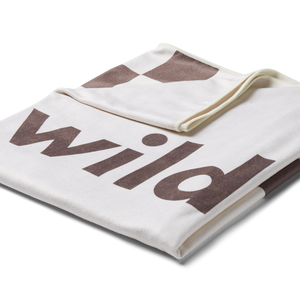 a 3/4 overhead image of a stone or white beach towel folded into a square with the top left corner turned down revealing the checkered racing pattern in brown with the word 'wild' printed in large brown letters on the front. 