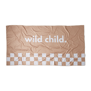 an overhead image of a beige microfiber beach towel with racing checkers along the bottom and the words 'wild child.' printed in large white letters across the middle on a flat surface showing the rectangular dimension of the towel. 