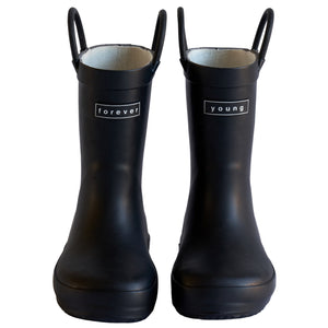 Front view of a pair of black colored rain boots for children. Showing pull handles for easy slip on and tags that say "Forever Young" on the front. 
