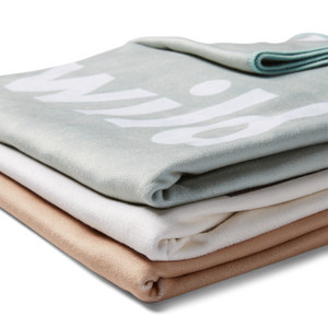 A stack of folded microfiber beach towels in beige, mint green, and stone colors, with the text 'Wild Child' visible on the gray towel.