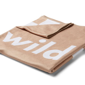 a 3/4 image of a beige beach towel folded into a square with the top left corner turned down revealing the checkered racing pattern in white with the word 'wild' printed in large white letters on the front. 