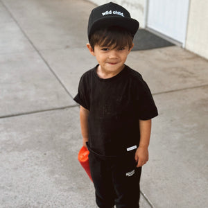 lifestyle image of boy in all black wearing a baseball cap embroidered with 'wild child.'