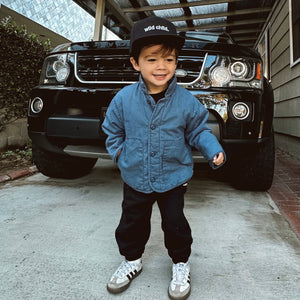 lifestyle image of boy standing in front of a Land Rover wearing a baseball cap embroidered with 'wild child.'