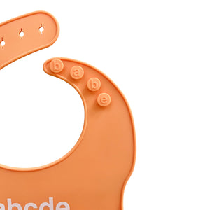 Close-up of an orange silicone bib with letter shapes 'babe' on a white background