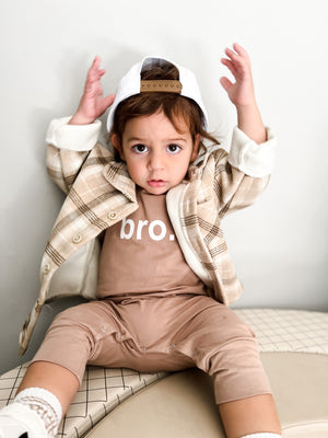 Baby sitting against wall throwing hands up in the air wearing brown 'bro.' baby romper. 
