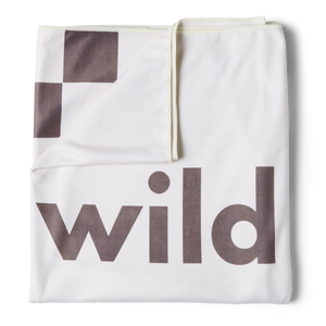 an overhead image of a stone or white beach towel folded into a square with the top left corner turned down revealing the checkered racing pattern in brown with the word 'wild' printed in large brown letters on the front. 
